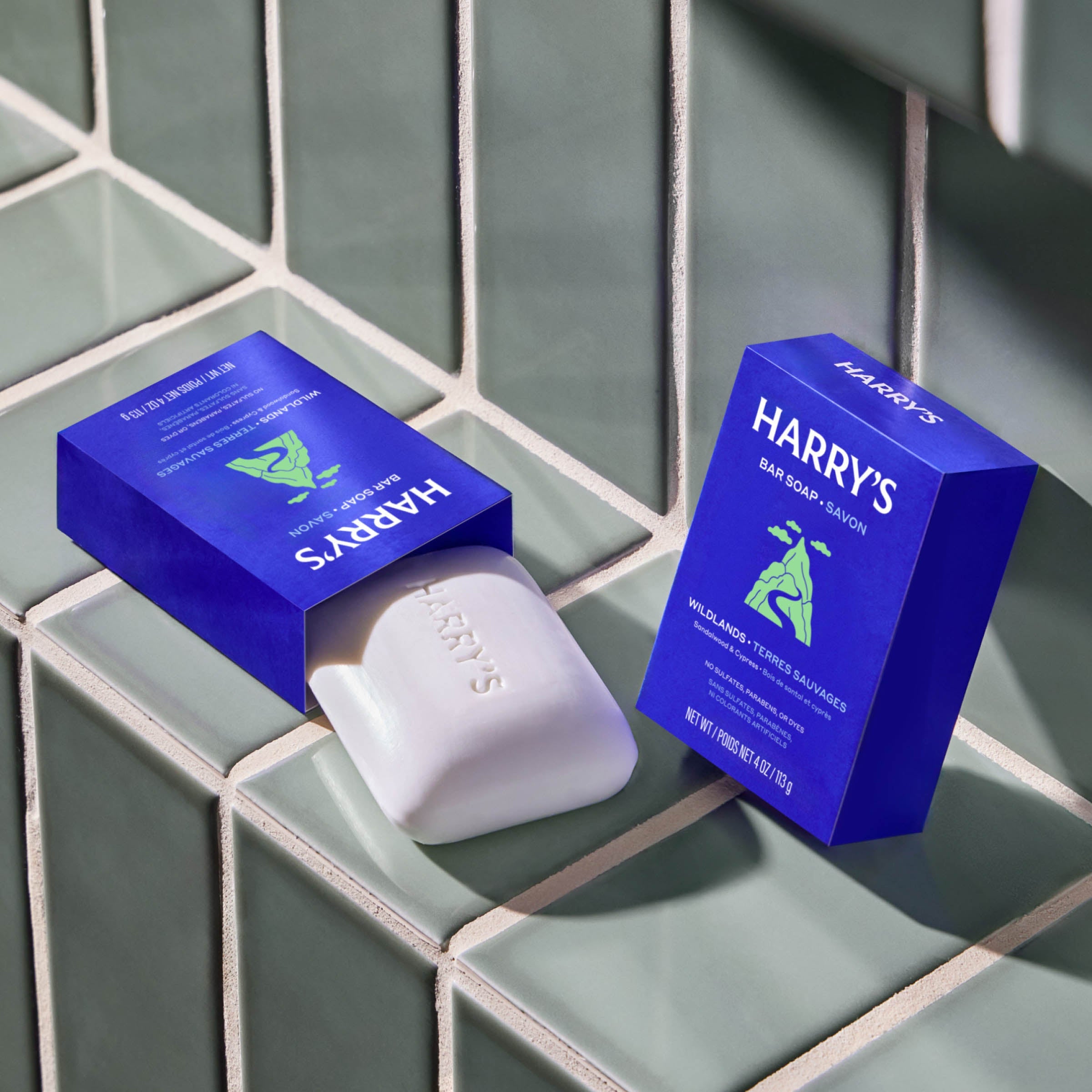 Two blue boxes of Harry's Soap with a white soap bar resting on a glossy green tiled surface.