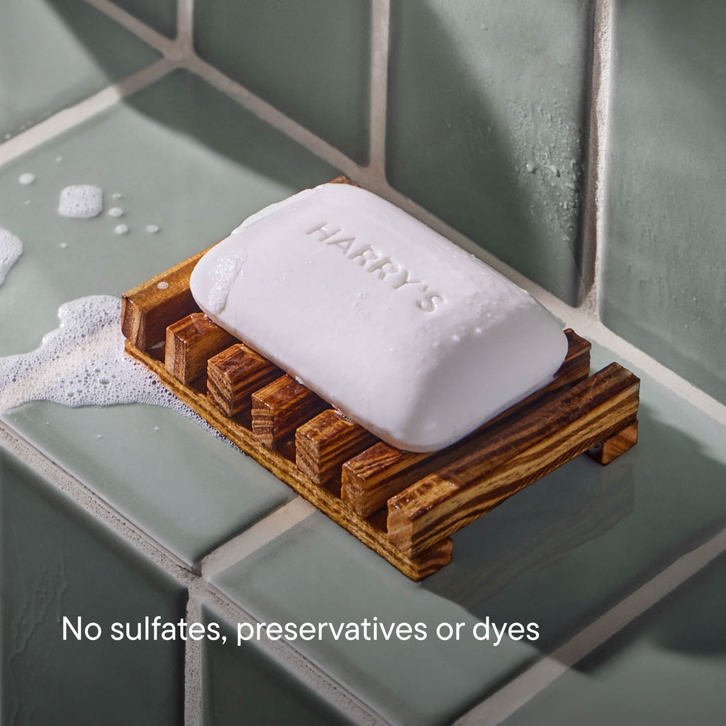 A white bar of soap labeled 'HARRY'S' rests on a wooden soap dish set on a tiled bathroom ledge with soap foam nearby. Text at the bottom reads 'No sulfates, preservatives or dyes'.