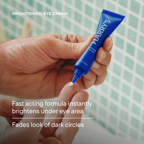 Hand holding a blue tube of Harry's brightening eye cream with text on a tiled wall background.