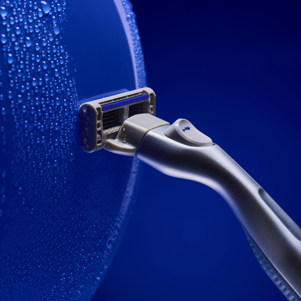 A close-up studio shot of a silver electric razor with a blue protective cover on the blade, positioned against a dark blue background with water droplets on a curved surface.