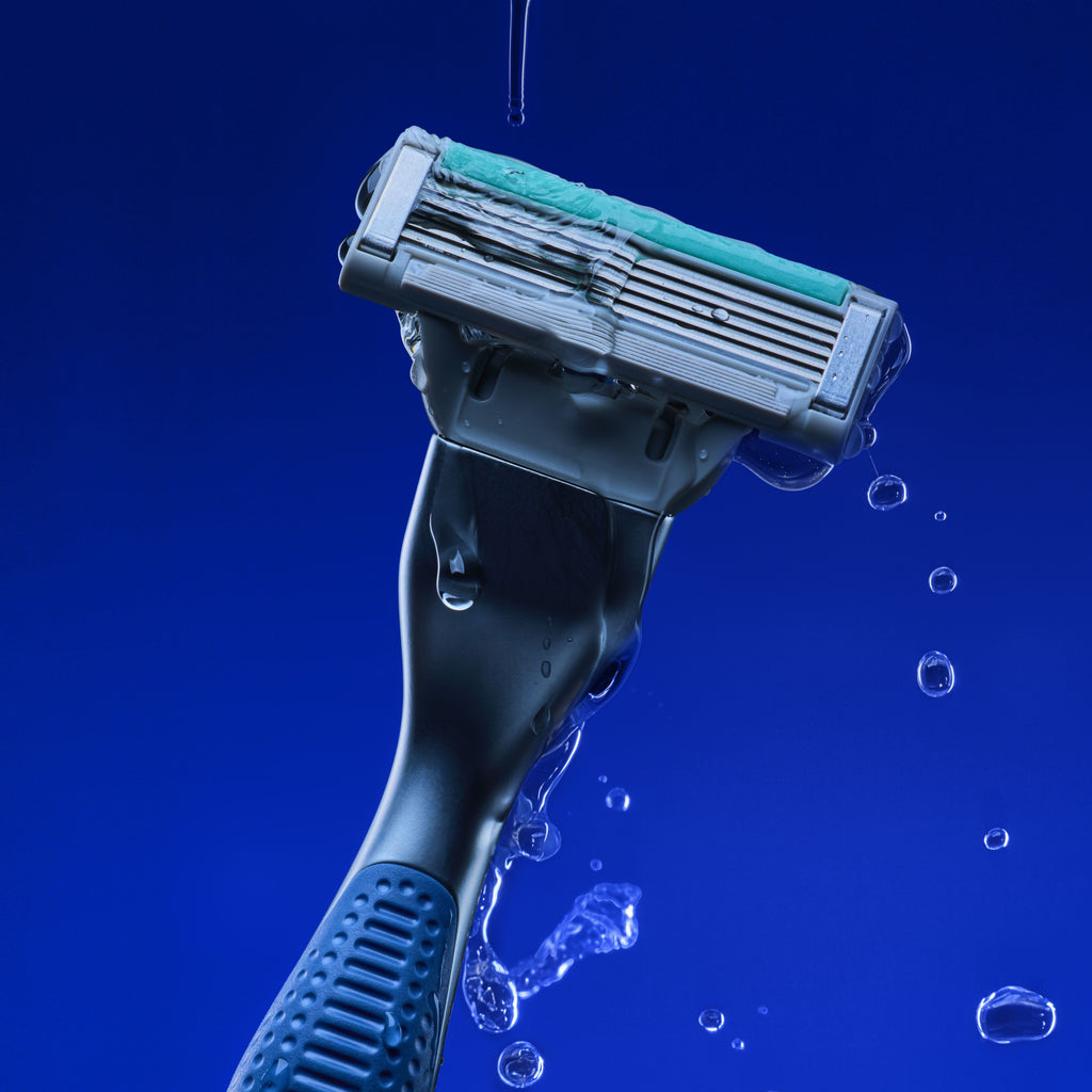 A close-up studio shot of a razor with a teal and gray head and a dark blue handle, shown with water droplets dripping off against a solid blue background.